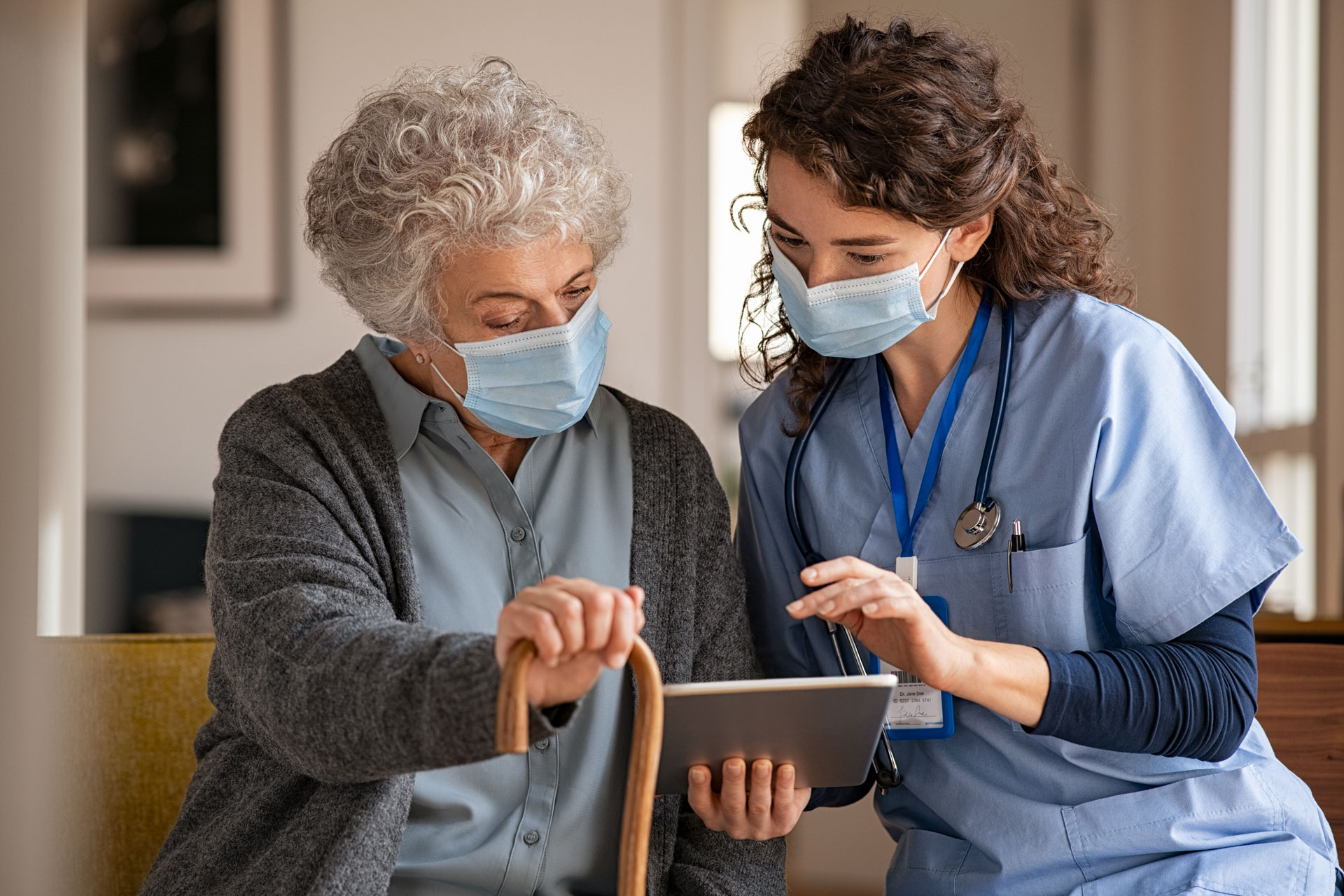 Young doctor and senior woman going through medical record on digital tablet during home visit wearing face protective mask.