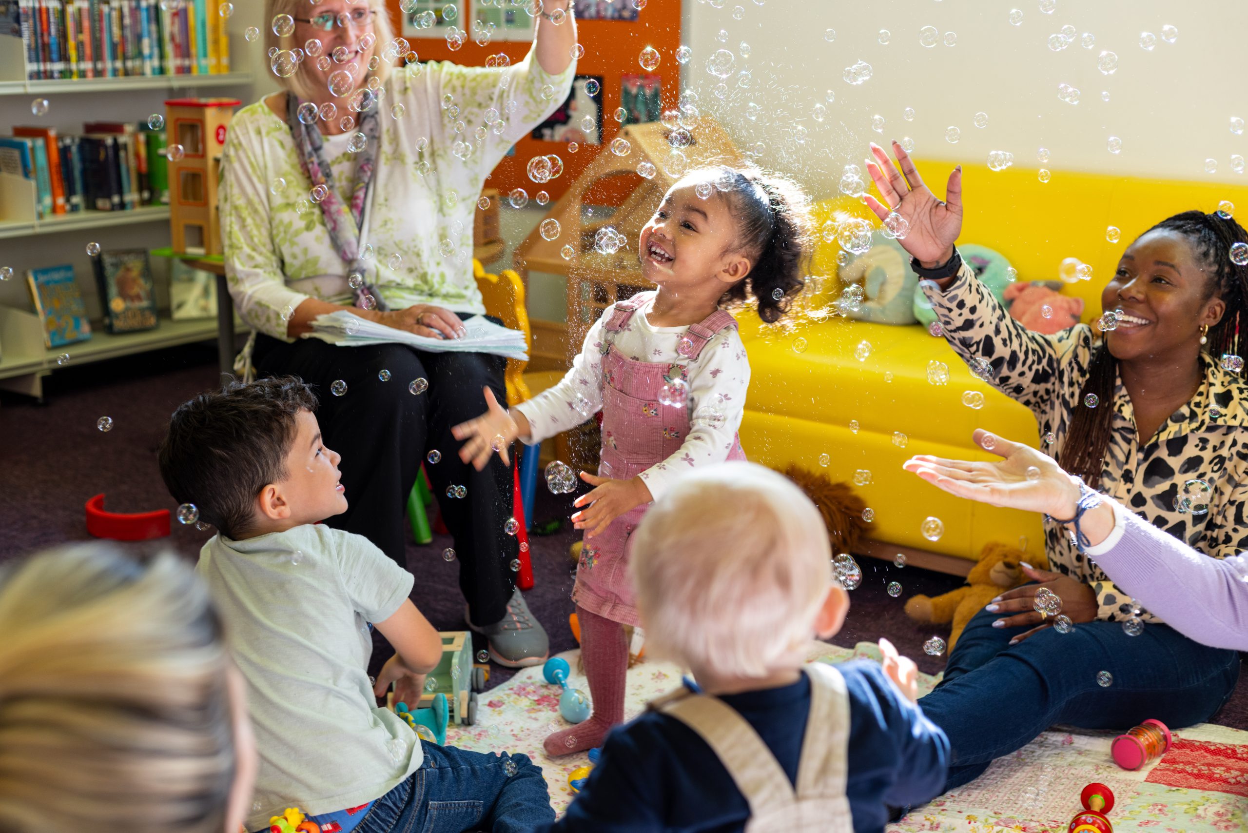 Medium shot of a group of adults and children gathered together in a community library in Newcastle-upon-Tyne, North East England. There is a teacher using a bubble machine while the group raise their arms joyfully.