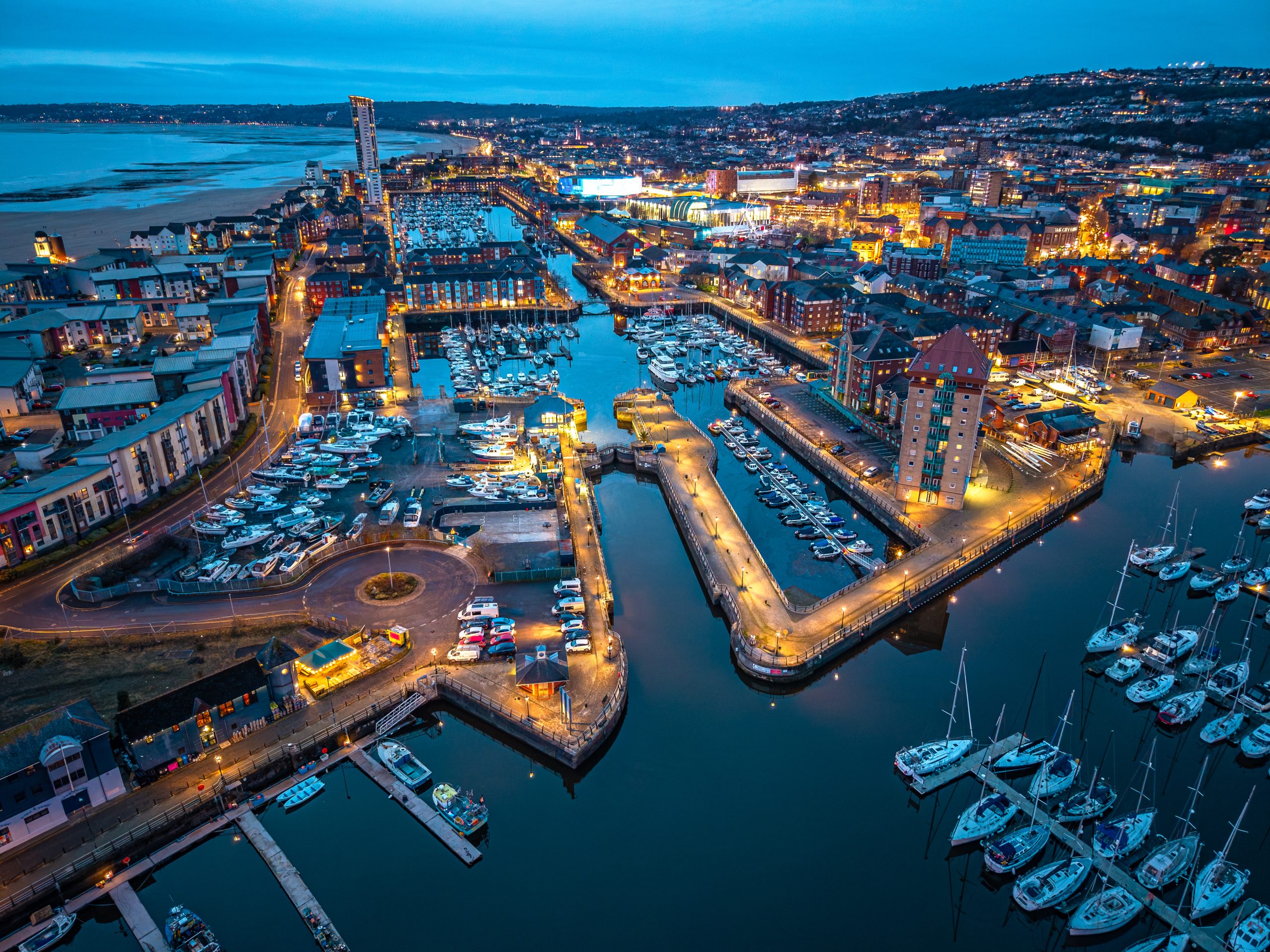 High angle aerial drone photograph of Swansea Marina at dusk (blue hour), showing illuminated city waterfront buildings, boats in the marina and Swansea Bay in the background, Swansea, Wales, United Kingdom.